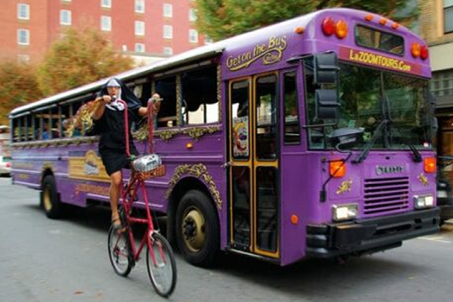 Large Purple Tour Bus with Man on Tall Bicycle riding beside
